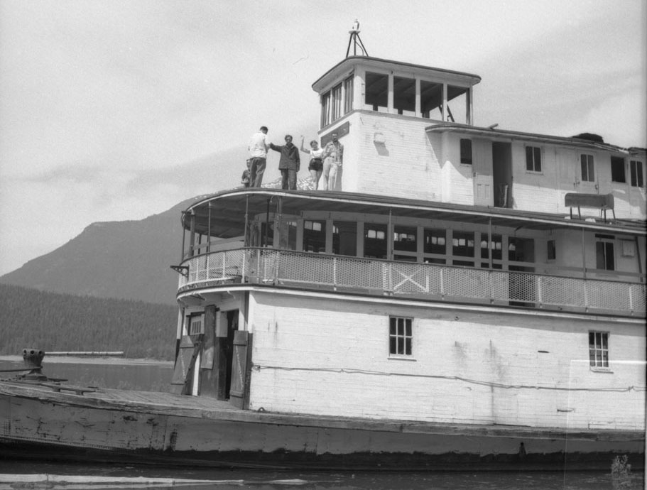 A black-and-white photograph of four people standing on top of a steamer boat. The photo cuts off the second half of the boat. A mountain and some water can be seen in the background on the left side.