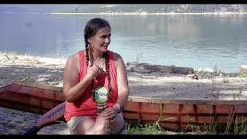 Shelly Boyd – a Sinixt woman with dark hair in two braids, wearing an orange tank-top. She is sitting on a log in front of a river with a canoe sitting behind her.