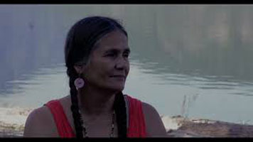 Shelly Boyd – a Sinixt woman with dark hair in two braids, wearing an orange tank-top. The image is a portrait view of Shelly with the water behind her.