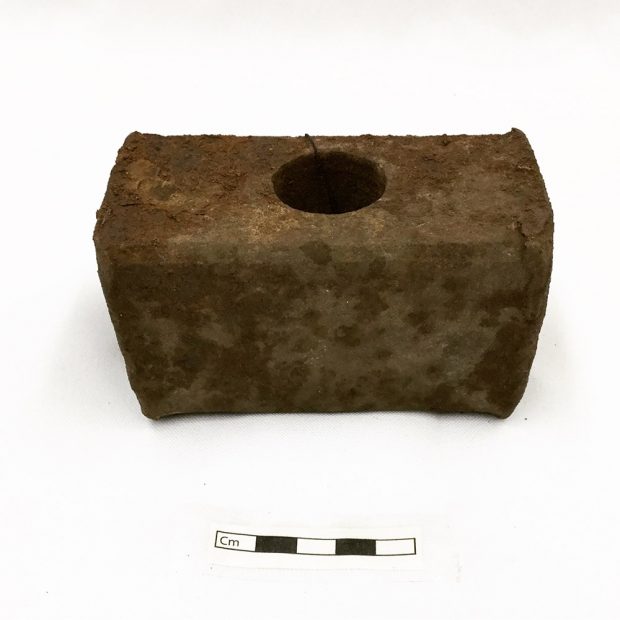 Colour photograph of a rectangular rusty metal sledgehammer head. There is a circular hole through its middle