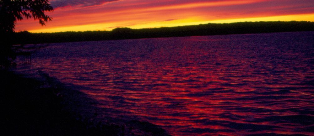 Colour photo of a body of water at sunset, sky displays yellow, orange, and purple hues, the water appears purplish.