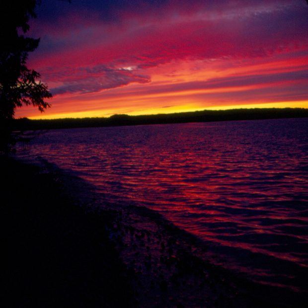 Colour photo of a body of water at sunset, sky displays yellow, orange, and purple hues, the water appears purplish.