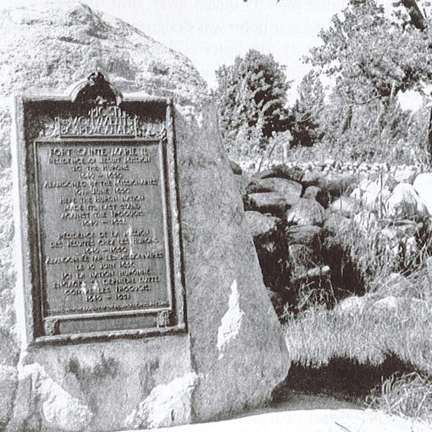 Black and White image of commemorative plaque affixed to a boulder with stone building foundations in the background