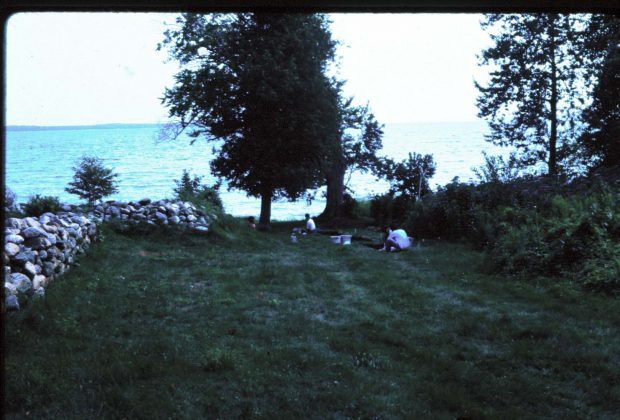 Image of low cobble walls arranged in an L shape on a grassy field with the lake behind.