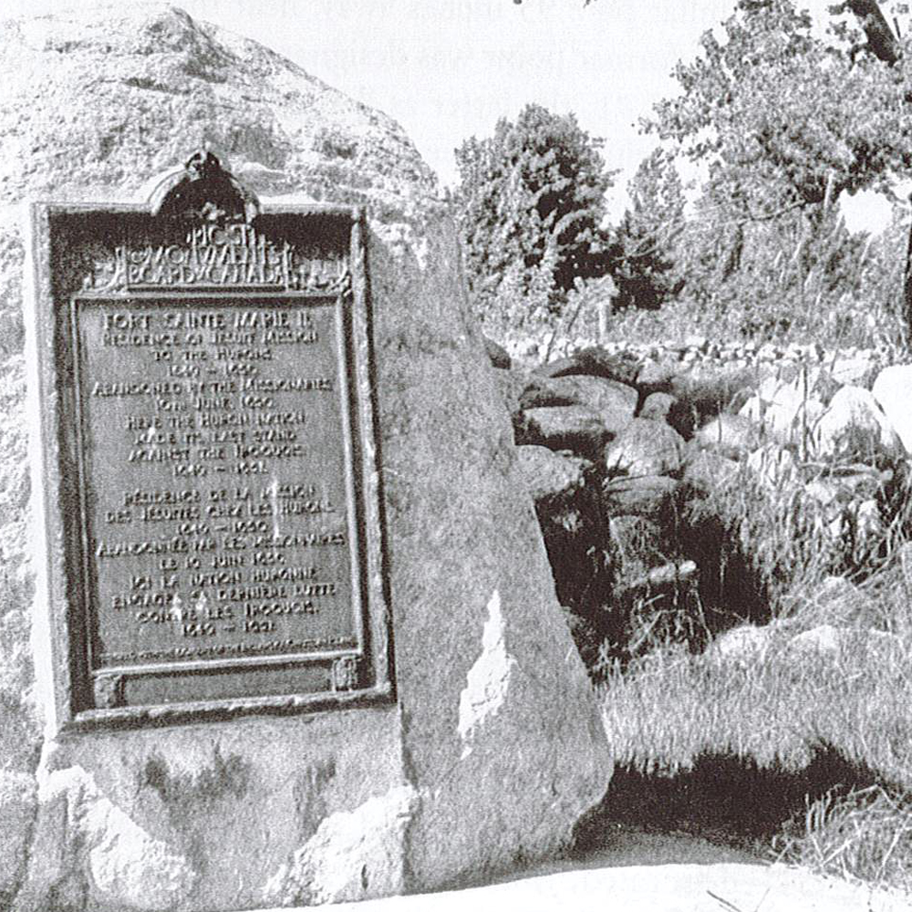 Image en noir et blanc d’une plaque commémorative fixée à un rocher avec des fondations en pierres en l’arrière plan.