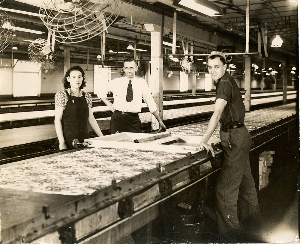 Three employees, a man in a white shirt and tie, a man in work clothes and a woman wearing an apron, pose in front of a long table in a factory.