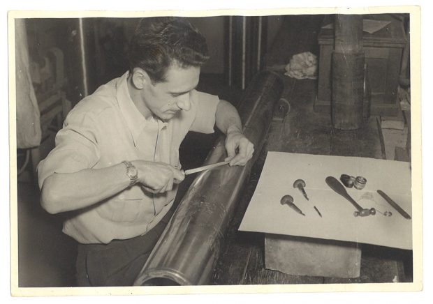 A workman bends over a roll of metal with small engraver's tools around it.