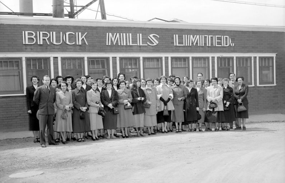 Un groupe de jeunes filles rassemblées devant l’entrée d’une usine sous une enseigne affichant Bruck Mills Limited
