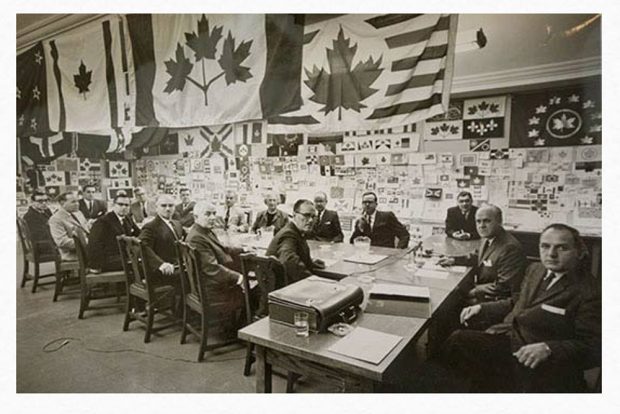 Black and white photo with 15 men seated at a long table with several flags hanging above them.