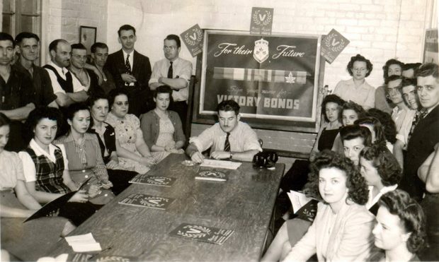A group of 28 employees around a table, women and men, pose during a promotional session for the purchase of victory bonds in 1945.