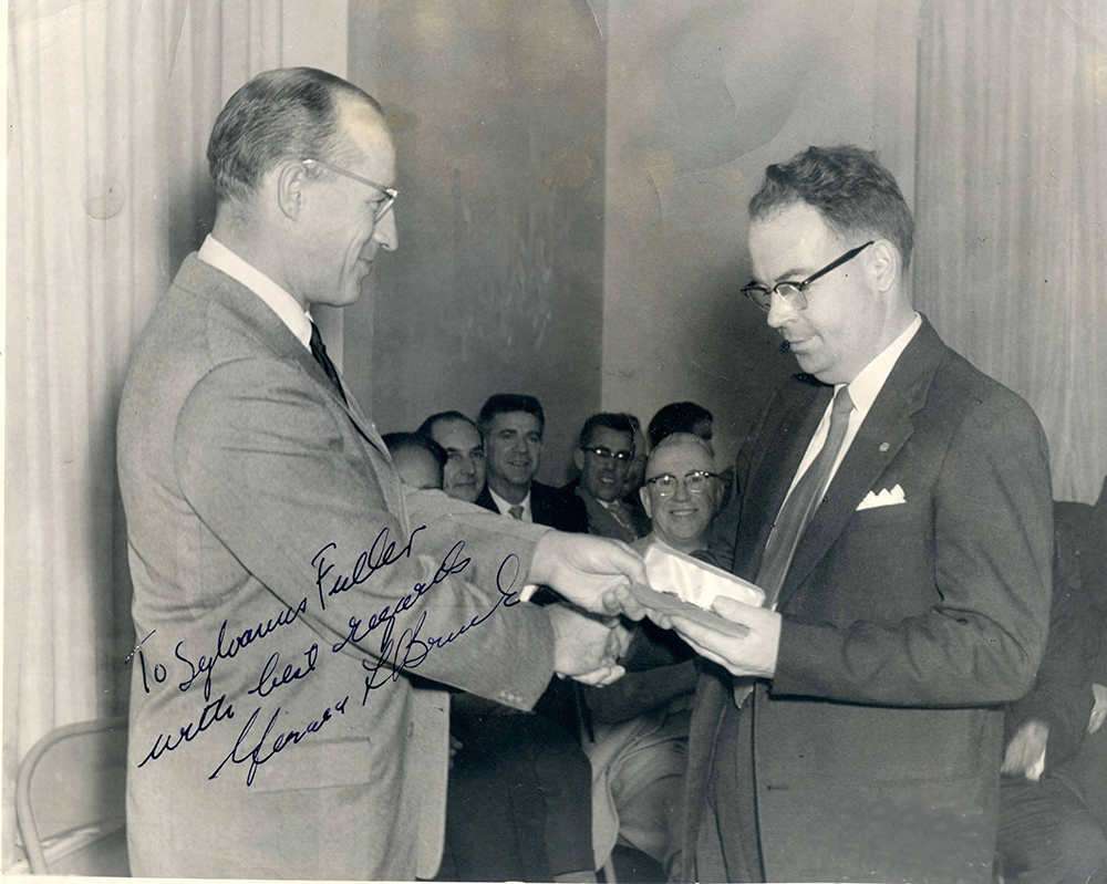 Autographed photo by Gerald Bruck showing the presentation of a watch to employee Sylvanus Fuller in front of guests