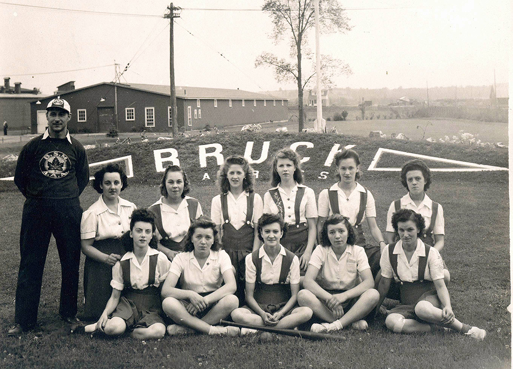 Women's ball team and their coach in two rows in front of an embankment marked "Bruck " with buildings in the background.