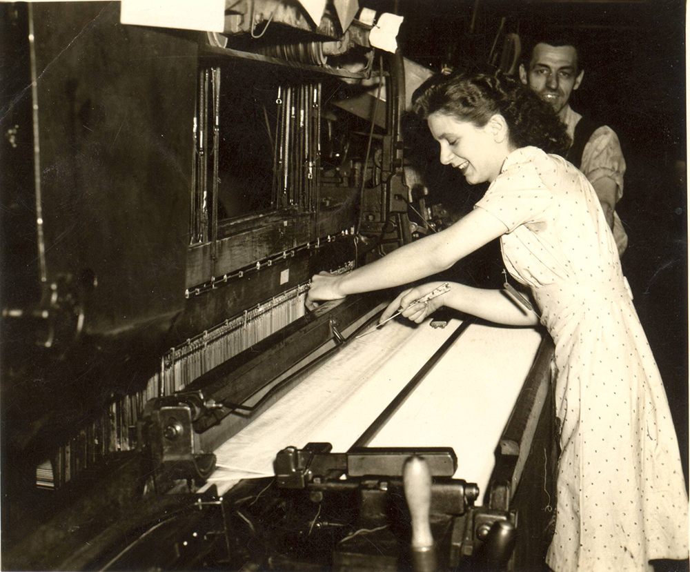A female worker stands in front of a loom and a man is standing behind her.