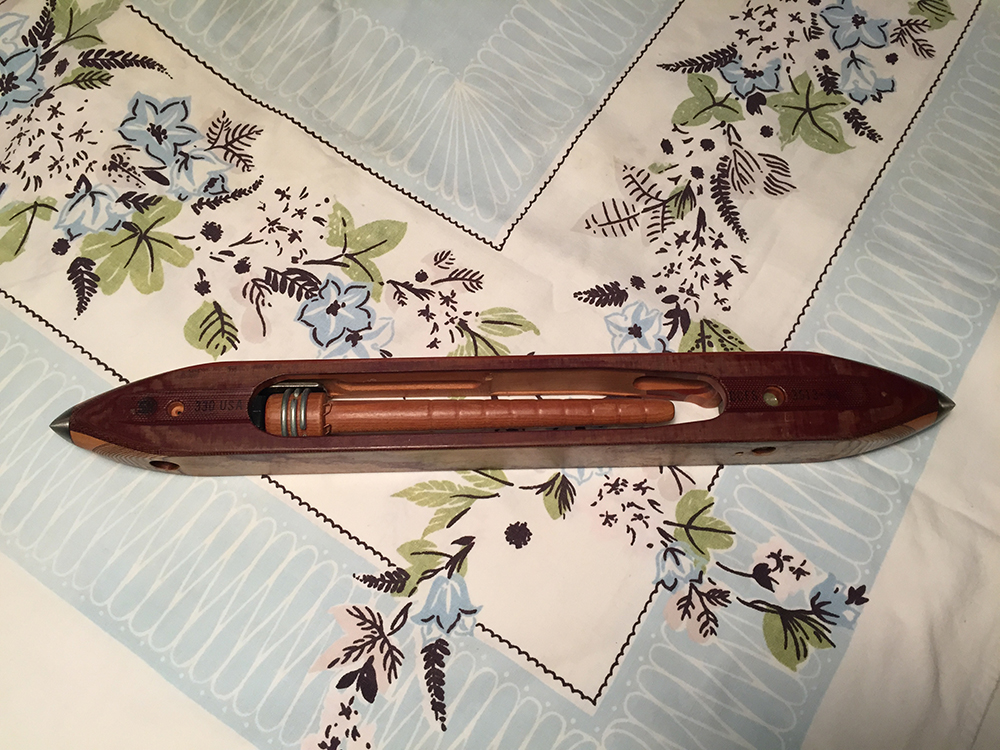 Section of a pale blue and white tablecloth with floral patterns. A wooden object called a shuttle is placed on the tablecloth