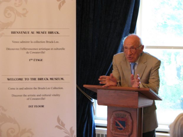 Elderly man (Gerald Bruck) in front of a window and leaning on a lectern with text on a banner on the left.
