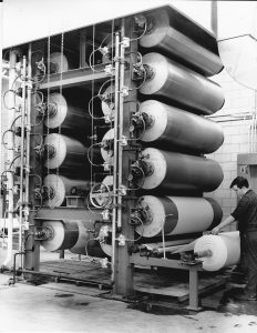 Black and white photo of a worker monitoring a device with several large stacked rolls of fabric.
