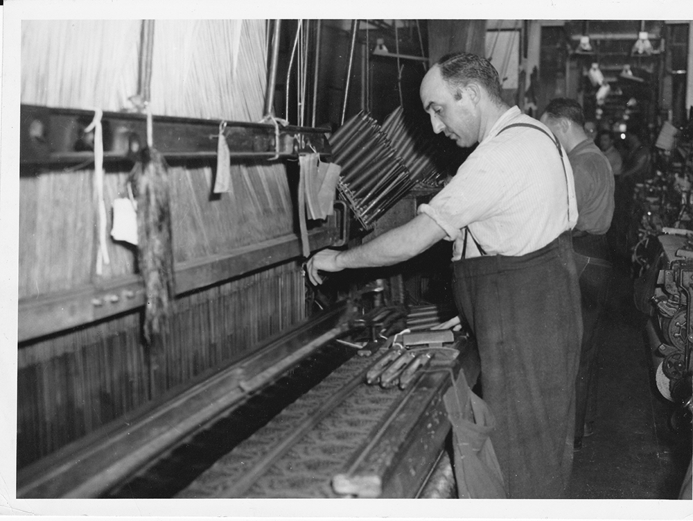 Black and white photo of a worker at his workstation in front of a loom.