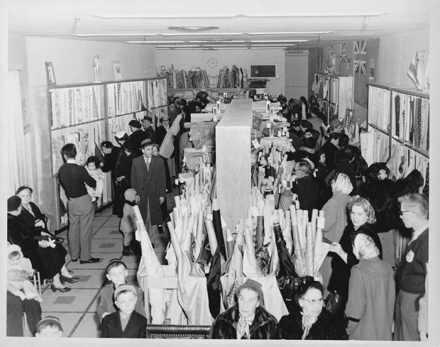 Black and white photo showing several customers around rolls of fabric in the Bruck's fabric store.