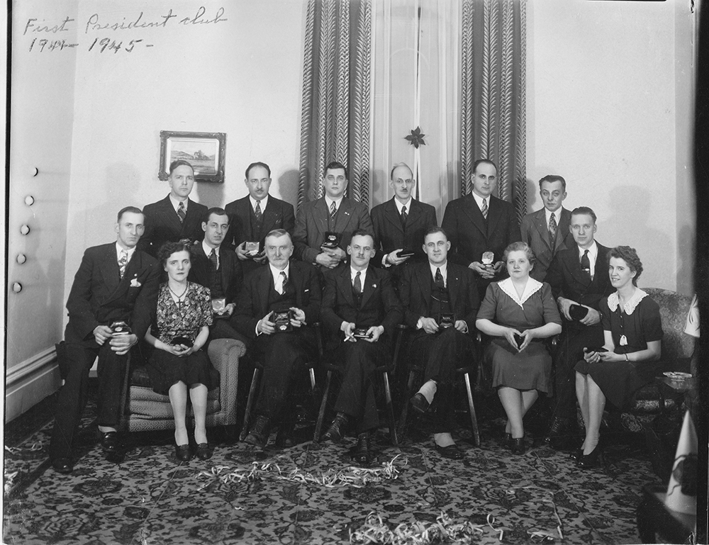 Black and white photo of members of the first President's Club pose in two rows with their gifts in their hands.