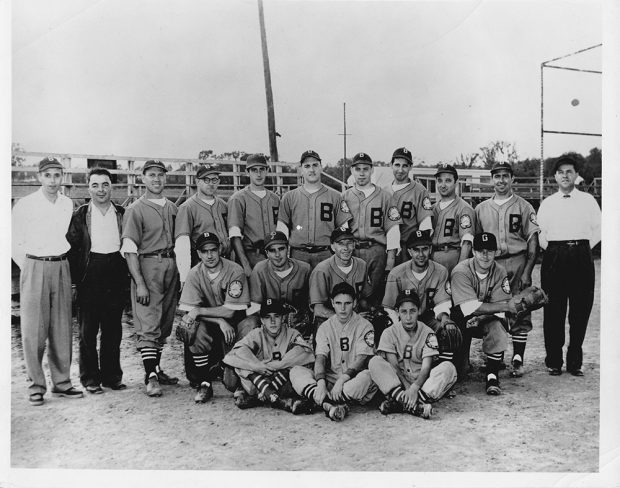 The Bruck baseball team in uniform on three rows with coaches, in front of a baseball field.
