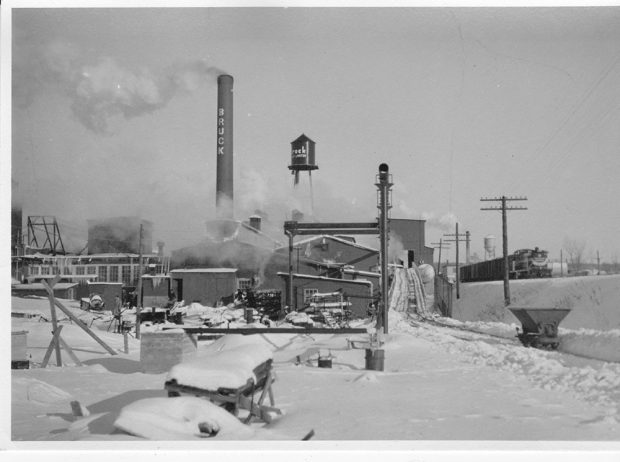 Black and white photo of the Bruck factory in winter with a train passing nearby on the right.