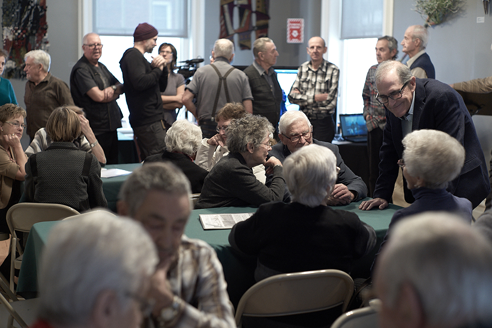 Color photo where elderly people are seated around several tables in the foreground and are talking. Other people are standing in the background talking in small groups in front of two windows and a screen.