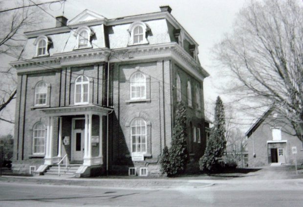 Black and white photo of a brick building with front columns and eight dormer windows. There is a smaller building to the right in the back with a pointed roof.