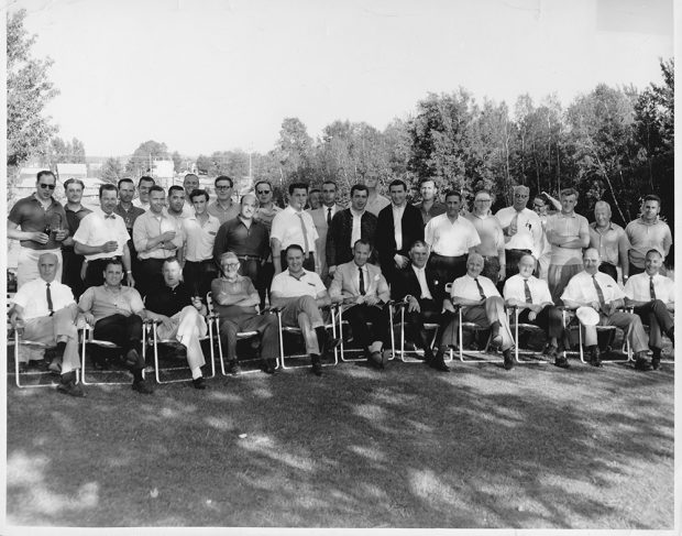 Three rows of men in shirts and ties on a golf course. Those in the first row are seated on lawn chairs.