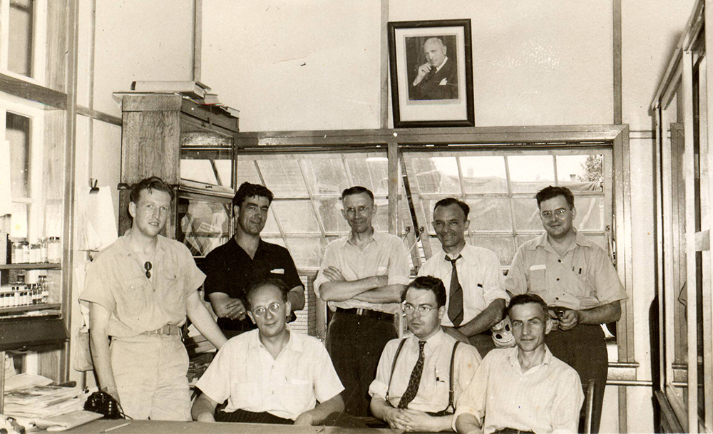 Black and white photo with eight employees sitting and standing in a factory office.