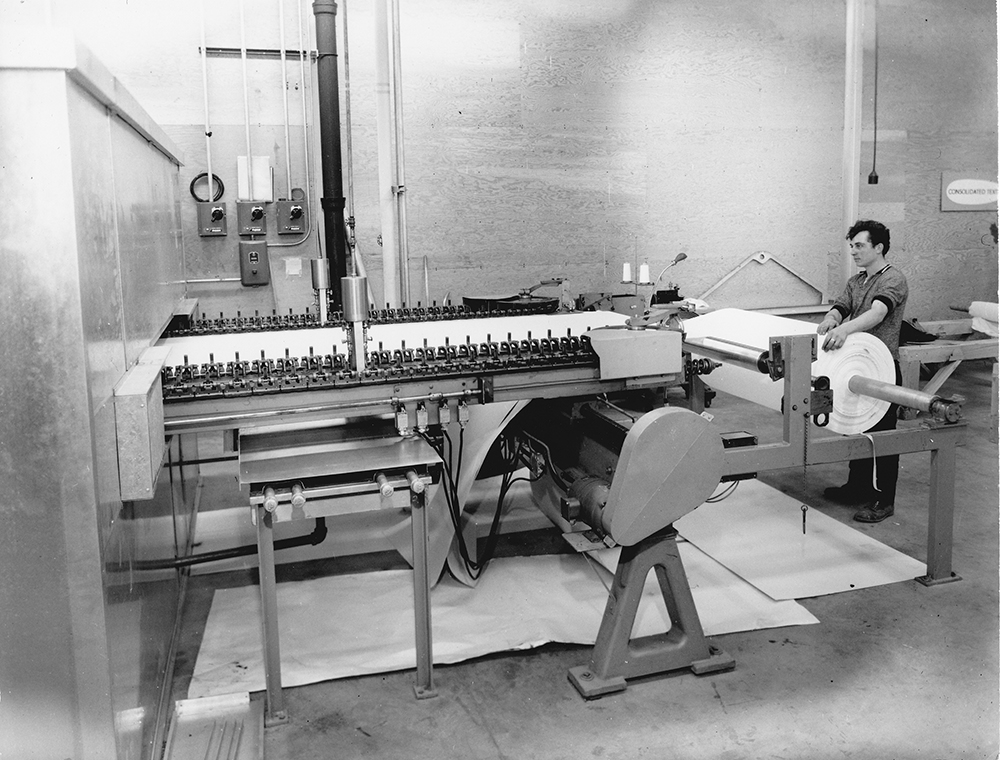 A worker monitors a roll of fabrics in front of a large machine.