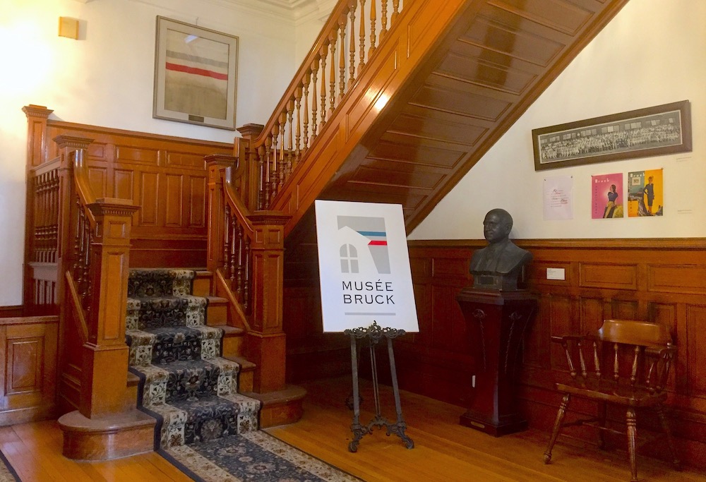 Photo of an entrance hall with a wooden staircase, a framed canvas, a poster on an easel and a bronze bust.