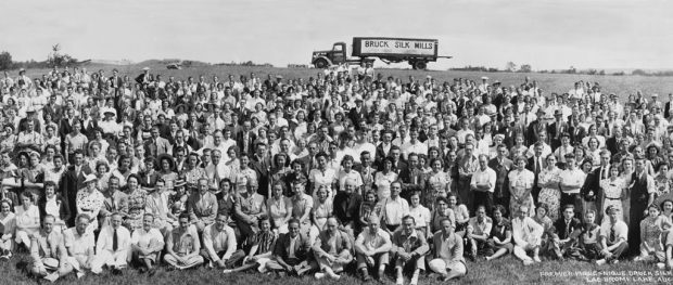 Black and white group photo with several rows of men and women who are sitting or standing in a field and looking at the camera. A parked truck appears in the distance, at the top of the photo.