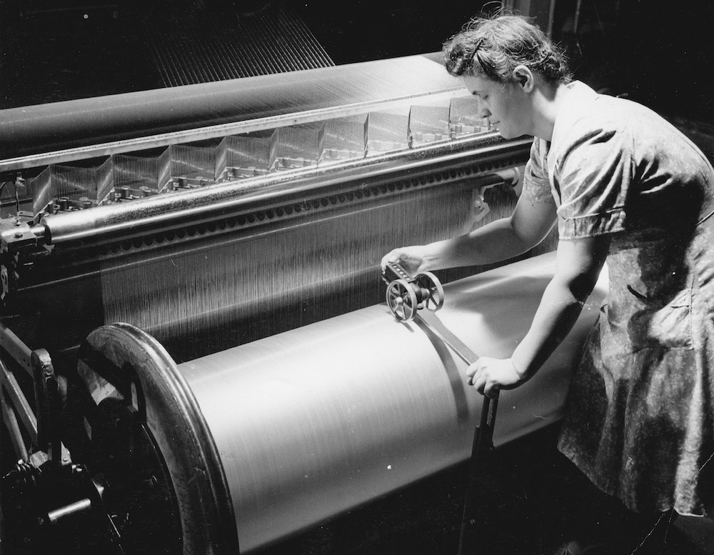 Black and white photo of a worker bent over a large roll of yarn connected to a machine