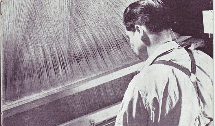 Black and white photo of a man seen from behind in front of a loom
