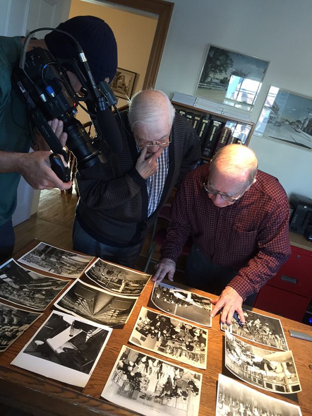 A cameraman films two elderly men looking at photographs placed on a table.