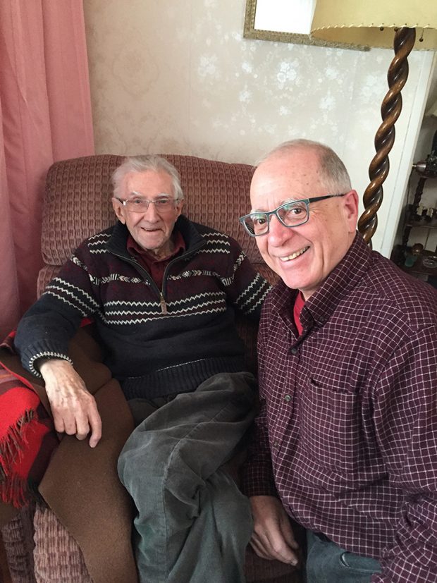 Elderly man seated in an armchair behind a smiling man in the foreground.