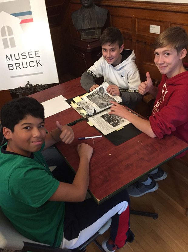 Three young boys seated in front of a school bag with a poster of the Bruck Museum logo and a bronze bust in the background.