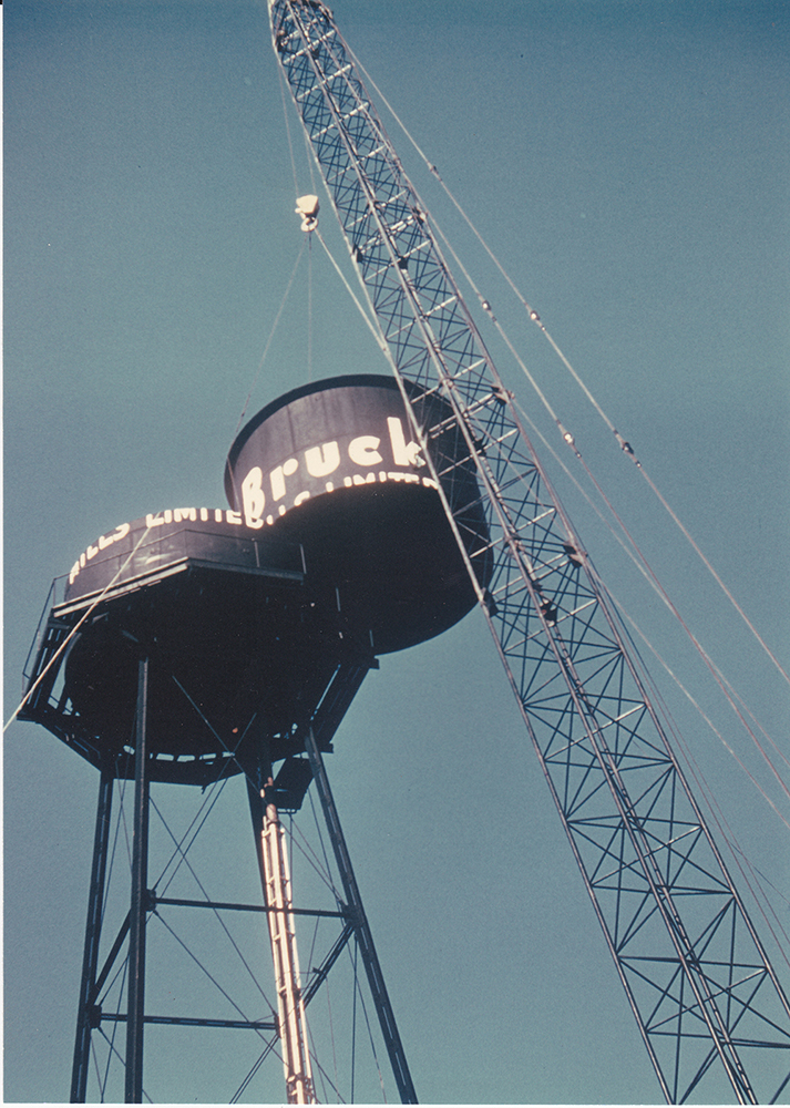 Huge crane dismantling a black wooden water tower with the inscription Bruck Mills Limited.
