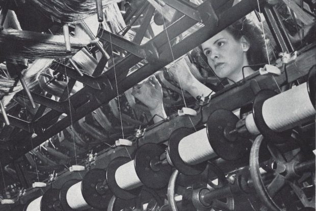 Black and white photo showing a worker looking at rows of spools of thread.