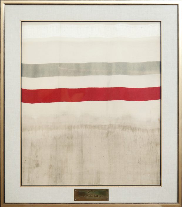 Frame displaying a piece of fabric with a red and gray stripe and a plaque identifying it as the first yard of woven silk in Canada.