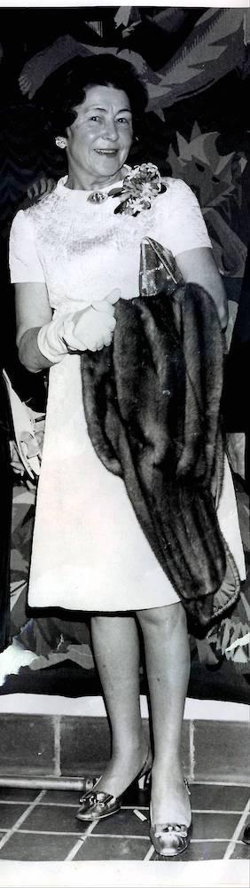 Black and white portrait of a smiling lady (Eugenie Sharp Lee) holding a small handbag and a fur coat under her arm.