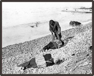 Black and white photo of a man examining two inanimate bodies on the beach.