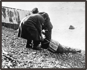 Photo en noir et blanc de deux hommes déplaçant un corps sur la plage.