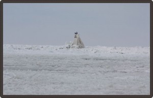 Photo of the ice-covered lake with a small building covered in snow above the ice in the centre.