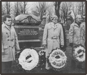 Black and white photo of three people in front of wreaths with tombs behind them.