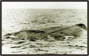 Photo en noir et blanc d’un navire chaviré flottant sur l’eau. On n’aperçoit qu’une portion de la coque du navire.