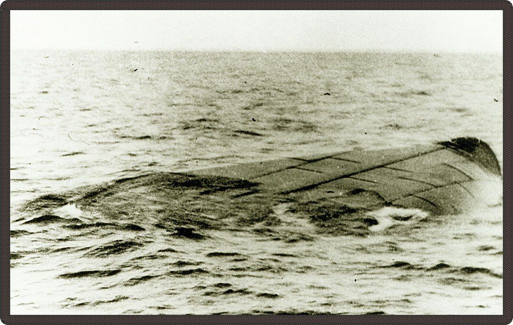 Black and white photo of an overturned vessel floating in the water with only a portion of the vessel's hull visible.