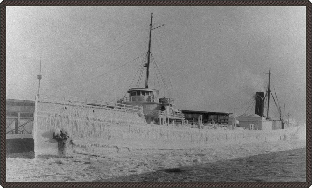 Black and white photo of a boat covered in snow and ice, stuck at harbour.