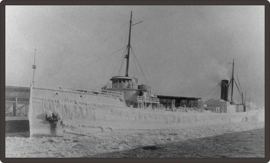 Photo en noir et blanc d’un bateau couvert de neige et de glace, immobilisé dans un port.