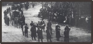Photo en noir et blanc montrant le cortège funèbre, précédé par des musiciens, défilant devant une foule.
